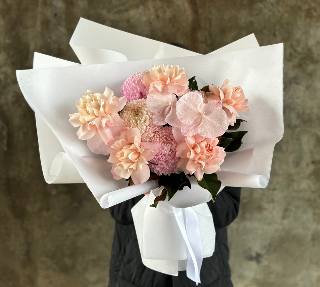 Close up image of Florist holding bouquet. A blush birthday bouquet of blush disbud, blush roses and blush phalaenopsis which is beautifully wrapped in KHF signature style. Florist wearing black and holding the guft bouquet against a concrete wall. 