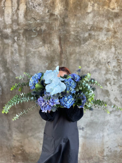 Florist holding a table hedge of blue seasonal flowers and grey blue foliages. Florist standing in front of a concrete wall.