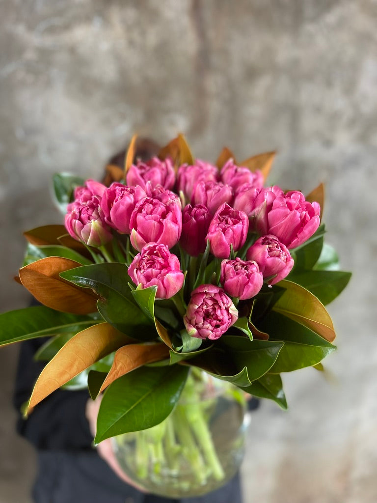 Close up image of Florist holding a clear tapered vase full of pink double tulips and magnolia foliage, in front of a concrete wall.