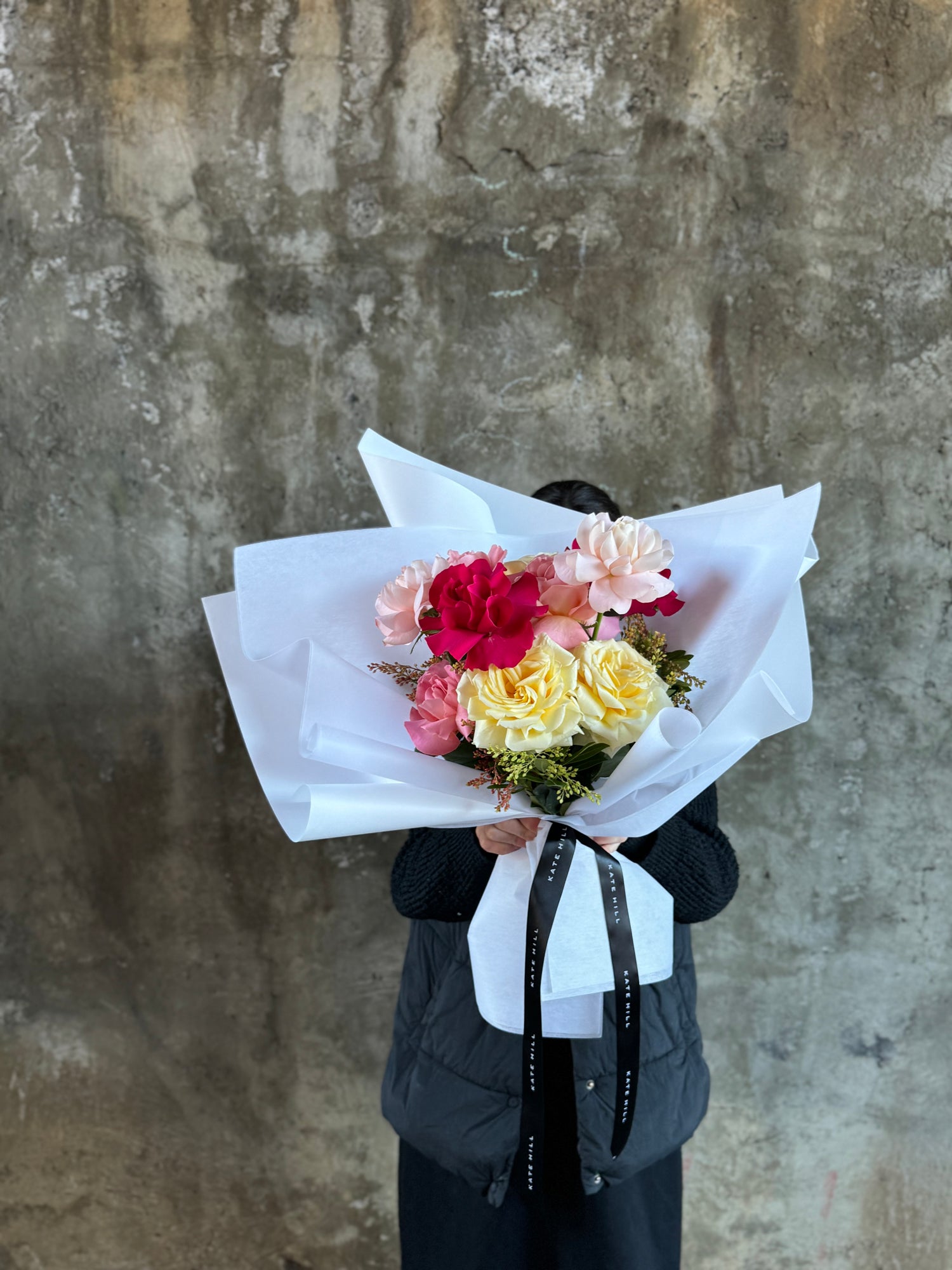 Wide image of Florist holding the bouquet of mixed coloured roses and standing against a concrete wall.
