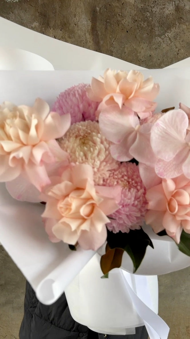 Video of the Chloe Flower Bouquet. A close up of a blush pink flower bouquet, beautifully wrapped in white signature paper. Florist holding the bouquet in front of a concrete wall.