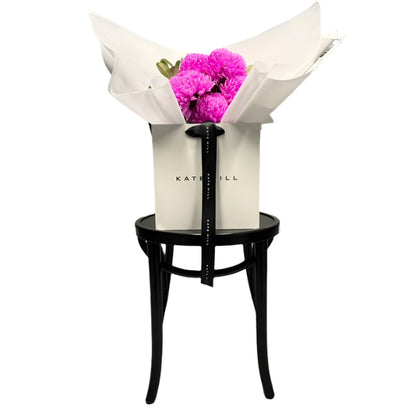 Bouquet of hot pink flowers in a white flower bag sitting on a black bentwood chair with a white wall background.