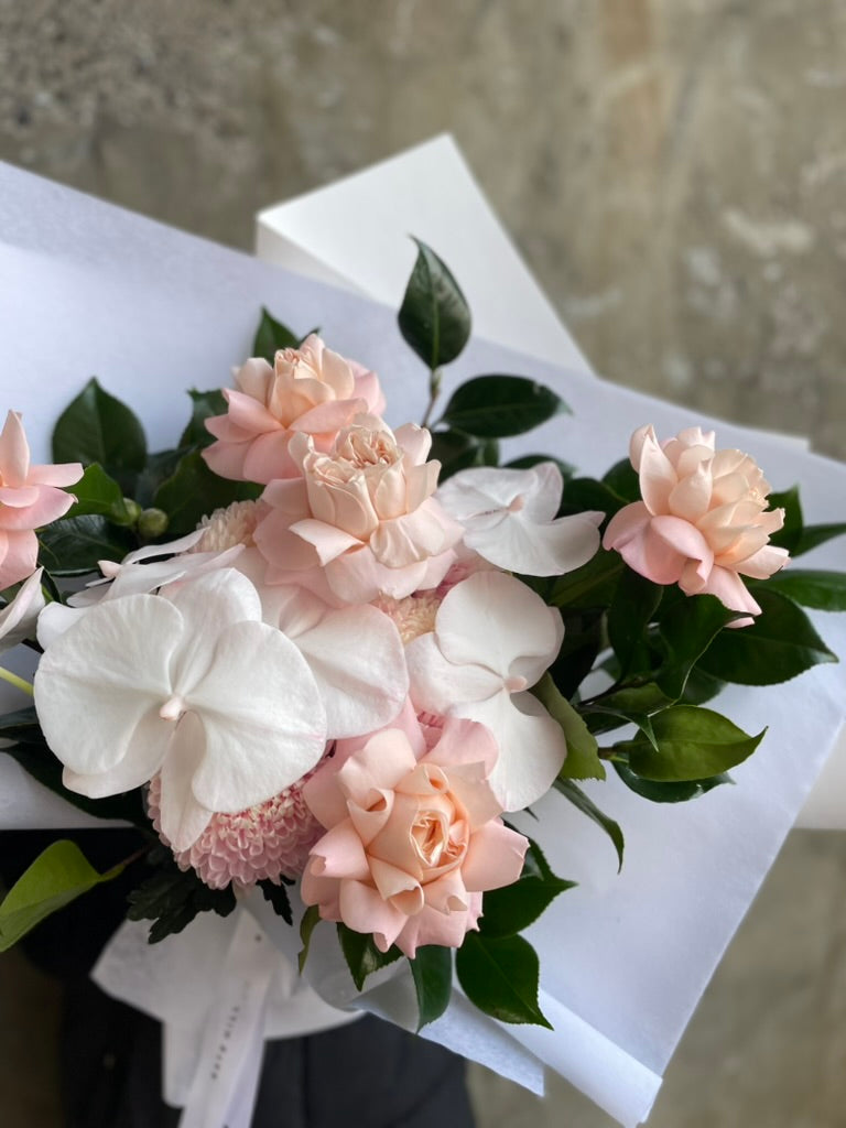 Close up image of the blush birthday bouquet wrapped in white paper, displayed against a concrete wall.