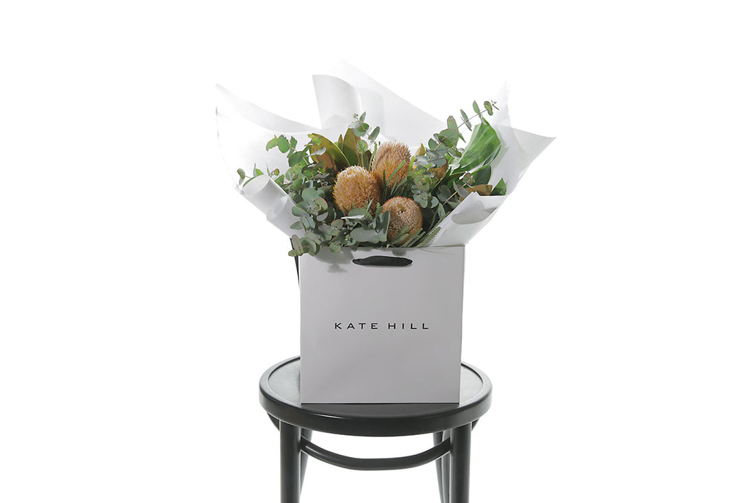 A australian native flower bouquet, beautifully wrapped in Kate Hill signature style (in flower bag) and sitting on a black bentwood chair, in front of a white wall.