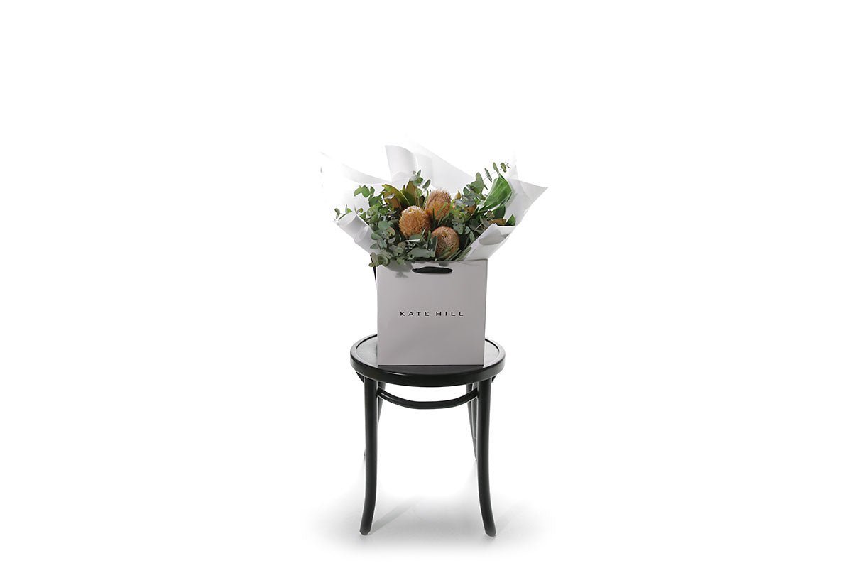 Wide image of an australian native flower bouquet, beautifully wrapped in Kate Hill signature style (in flower bag) and sitting on a black bentwood chair, in front of a white wall.