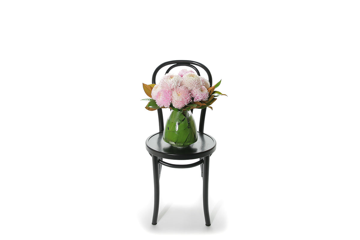 Wide Image of A mushroom vase, lined with green monsteria leaves, displying pink blush disbuds and a hint of foliage. Vase deisign sitting on a black bentwood chair with a white background.