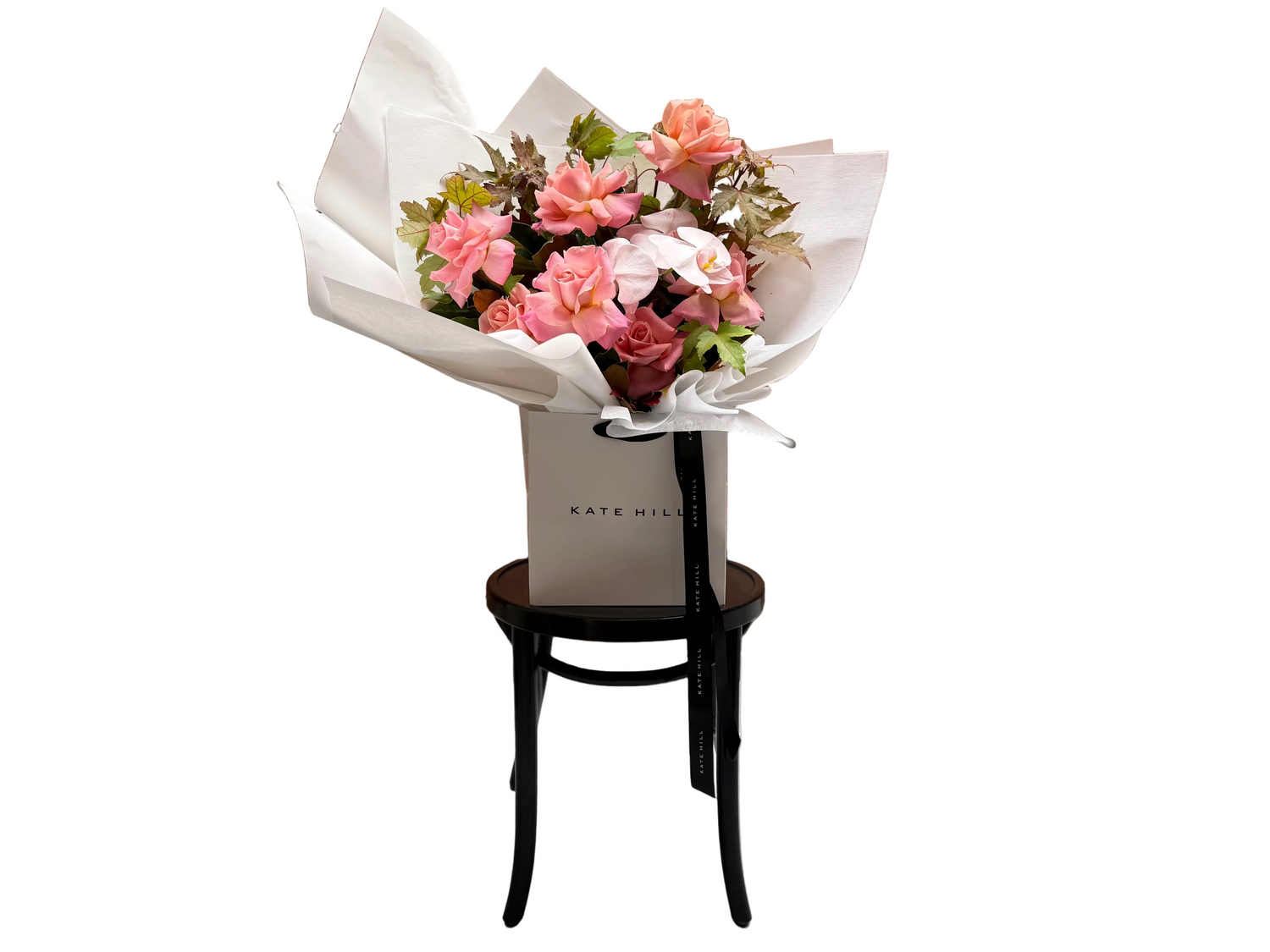 Our April Birthday Bouquet sitting on a black bentwood chair in front of a white wall. Bouquet in Flower Bag features watermelon roses, blush orchids and textural brown autumn maple and magnolia foliage.