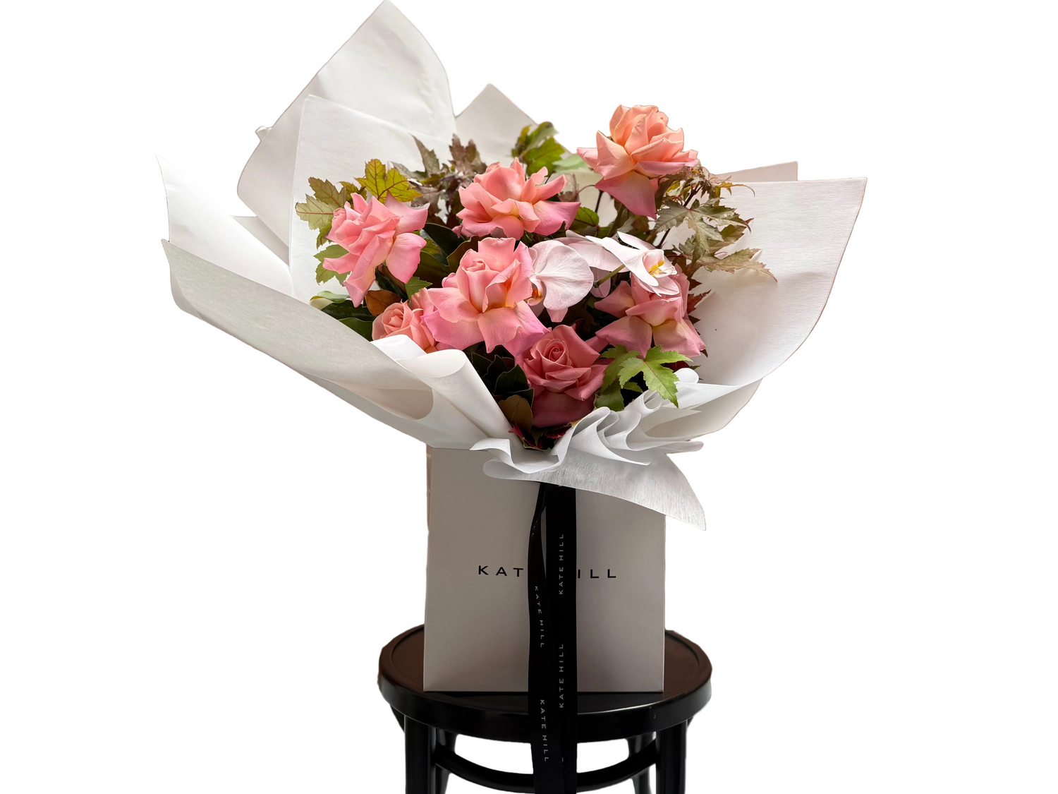 A close up of our April Birthday Bouquet sitting on a black bentwood chair in front of a white wall. Bouquet in Flower Bag features watermelon roses blush orchids and textural brown autumn maple and magnolia foliage.