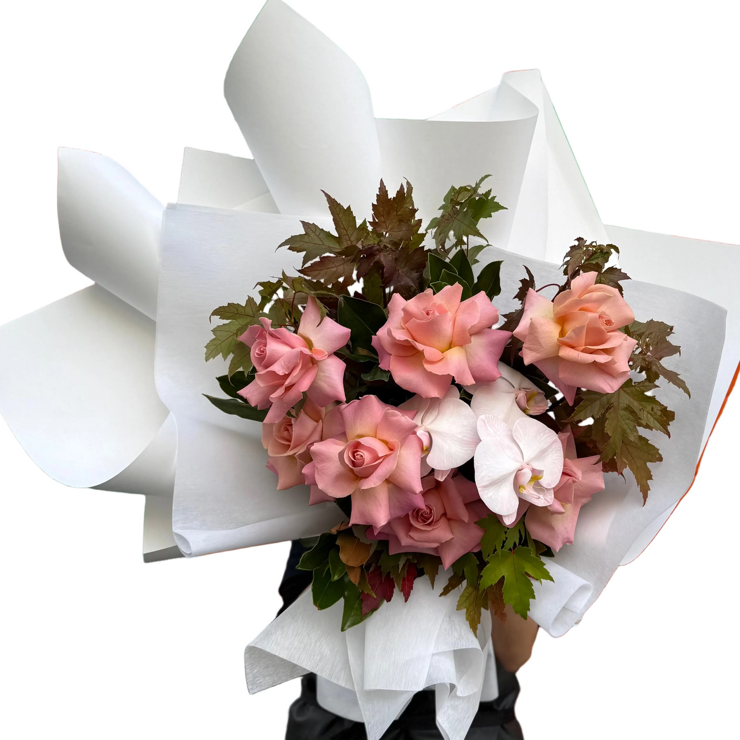 Our April Birthday Bouquet being held by a florist standing in front of a white wall. Bouquet features watermelon roses blush orchids and textural brown autumn maple and magnolia foliage.