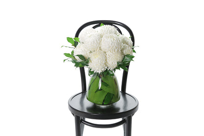 ARLO Tapered Vase Flower Design features white disbuds, a hint of foliage and is displayed into a leaf lined mushroom vase. Vase Design is sitting on a black bentwood chair with a white background.