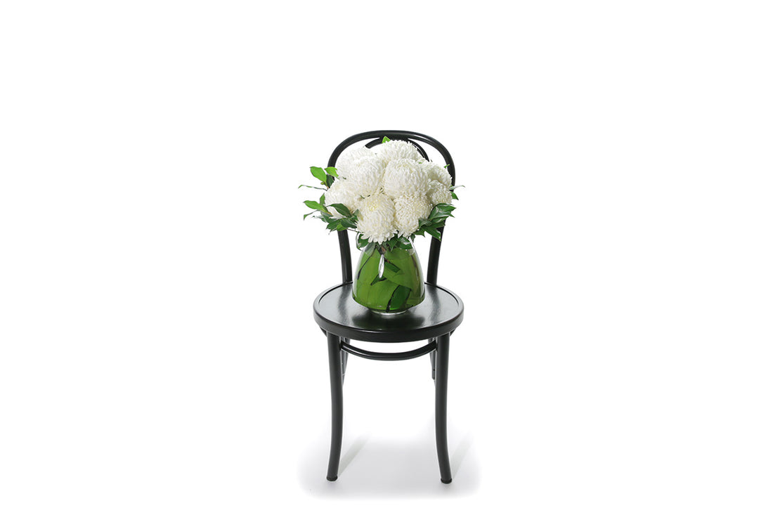 Wide image of ARLO Tapered Vase Flower Design features white disbuds, a hint of foliage and is displayed into a leaf lined mushroom vase. Vase Design is sitting on a black bentwood chair with a white background.