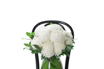 Close up image of ARLO Tapered Vase Flower Design features white disbuds, a hint of foliage and is displayed into a leaf lined mushroom vase. Vase Design is sitting on a black bentwood chair with a white background.