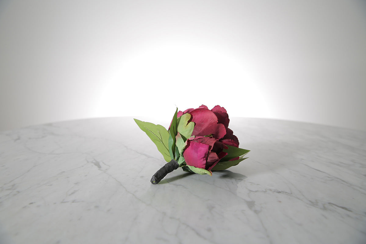 Kate Hill Flowers – artificial burgundy peony rose buttonhole flower with green leaf backing and pearl pin on marble table setting. 