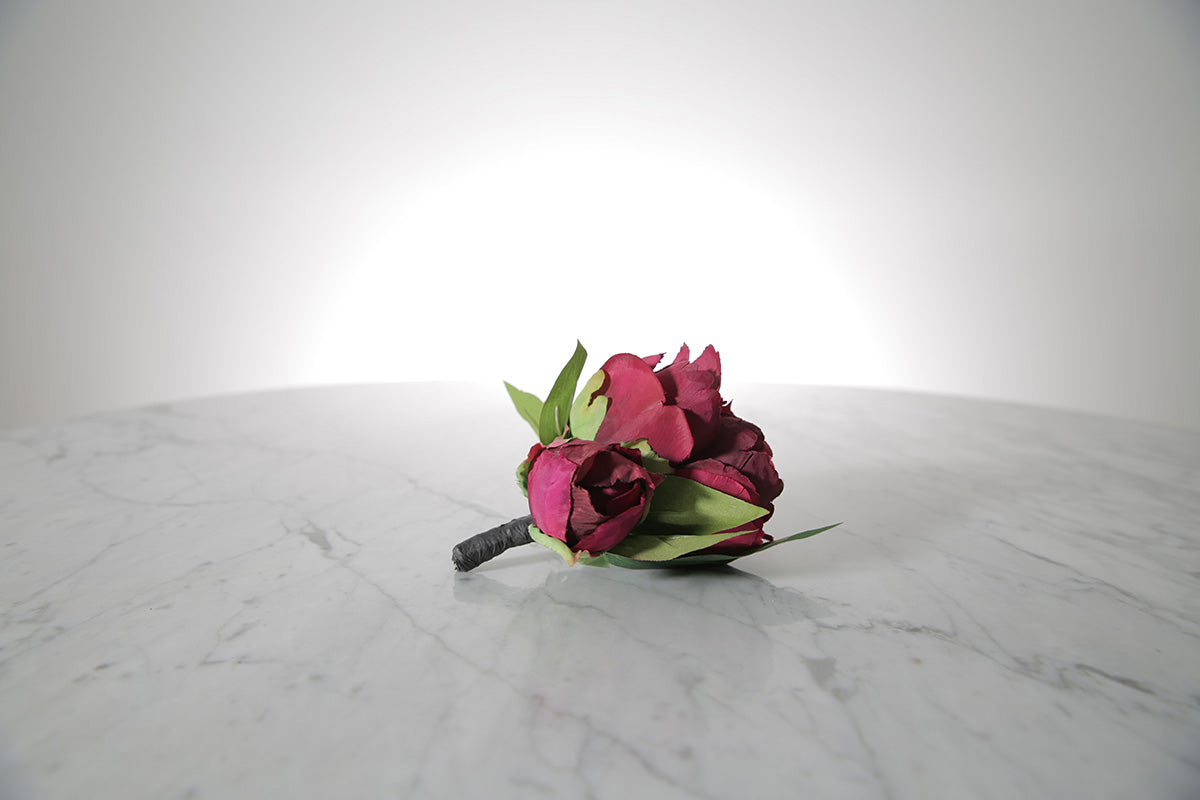 Kate Hill Flowers – artificial burgundy peony rose buttonhole flower with green leaf backing and pearl pin on marble table setting. 