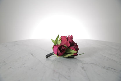 Kate Hill Flowers – artificial burgundy peony rose buttonhole flower with green leaf backing and pearl pin on marble table setting. 