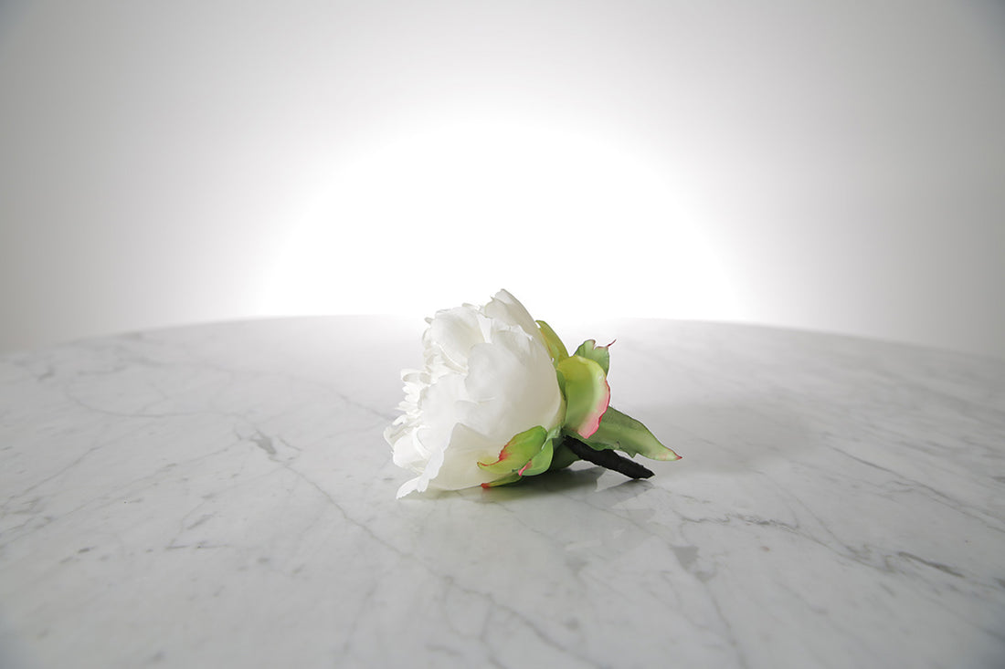 Kate Hill Flowers – artificial white peony rose buttonhole flower with green leaf backing and pearl pin set on a marble table top.