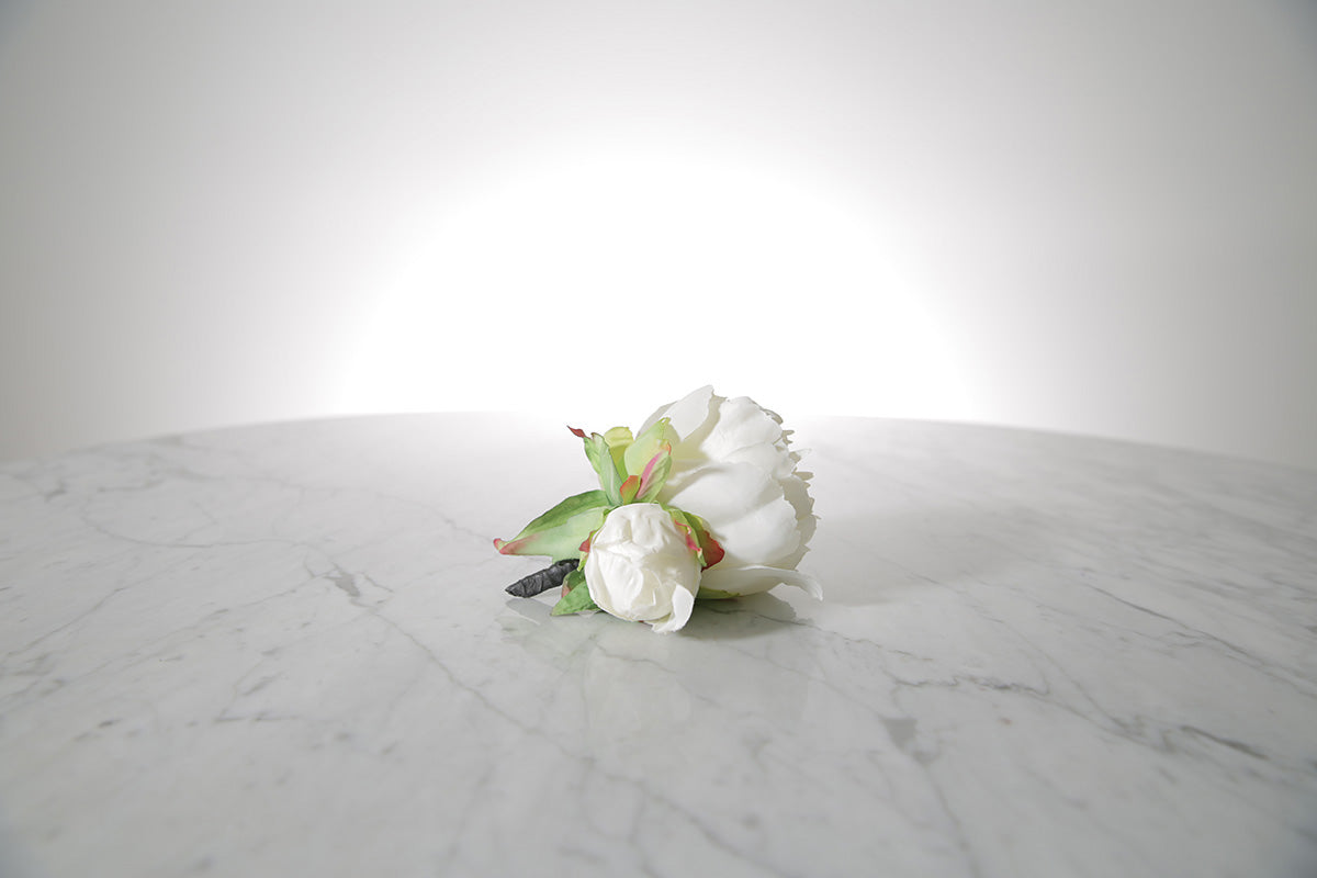 Kate Hill Flowers – artificial white peony rose buttonhole flower with green leaf backing and pearl pin set on a marble table top.