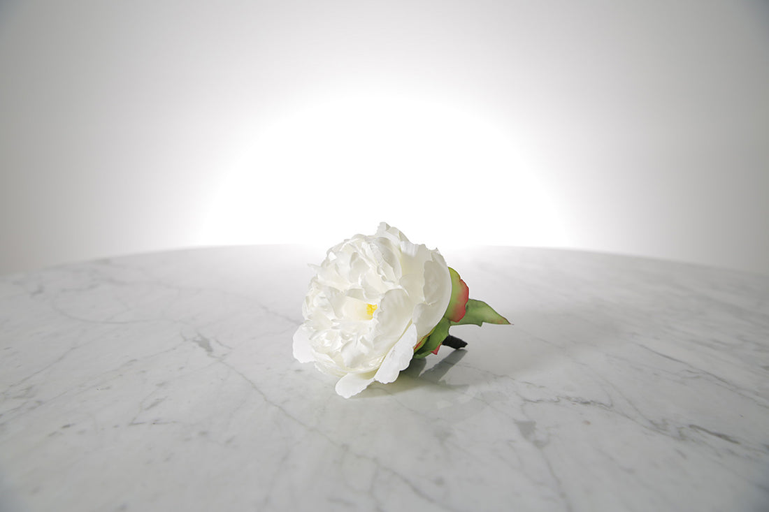Kate Hill Flowers – artificial white peony rose buttonhole flower with green leaf backing and pearl pin set on a marble table top.