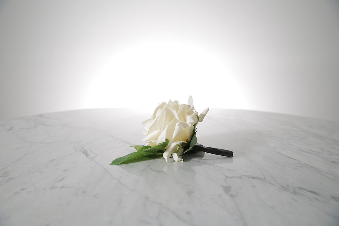 Kate Hill Flowers – artificial white rose buttonhole flower with green leaf backing, black-taped stem and pearl pin on a marble table setting.