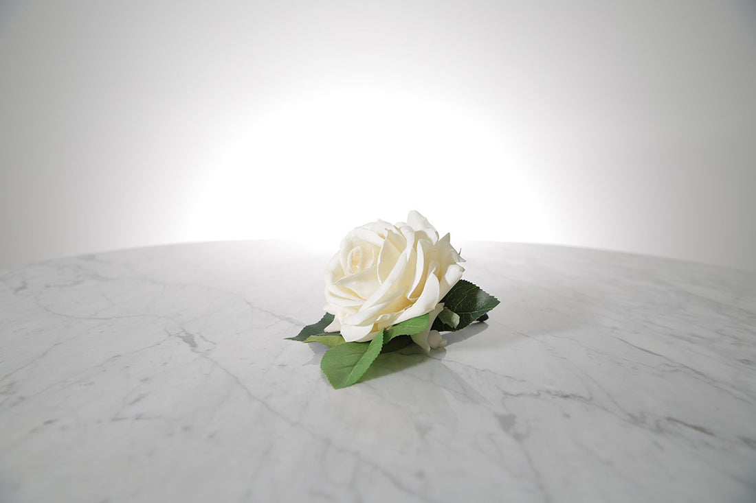 Kate Hill Flowers – artificial white rose buttonhole flower with green leaf backing, black-taped stem and pearl pin on a marble table setting.