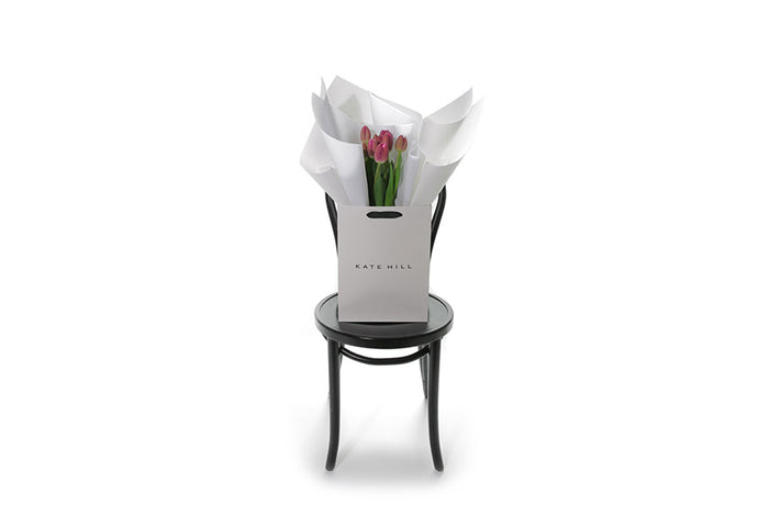 A bunch of 10 pink tulips wrapped beautifully in white signature wrapping paper and bouquet is placed into Kate Hill gift bag for presentation. Bag of pink tulips sitting on a black bentwood chair for photograph. Bouquet stands approximately 45cm tall on chair.