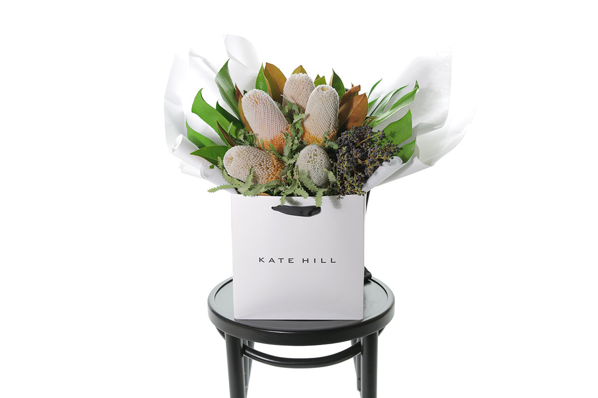 A seasonal native bouquet pictured with banksias and seasonal foliage. Bouquet sitting on a black bentwood chair with white background.