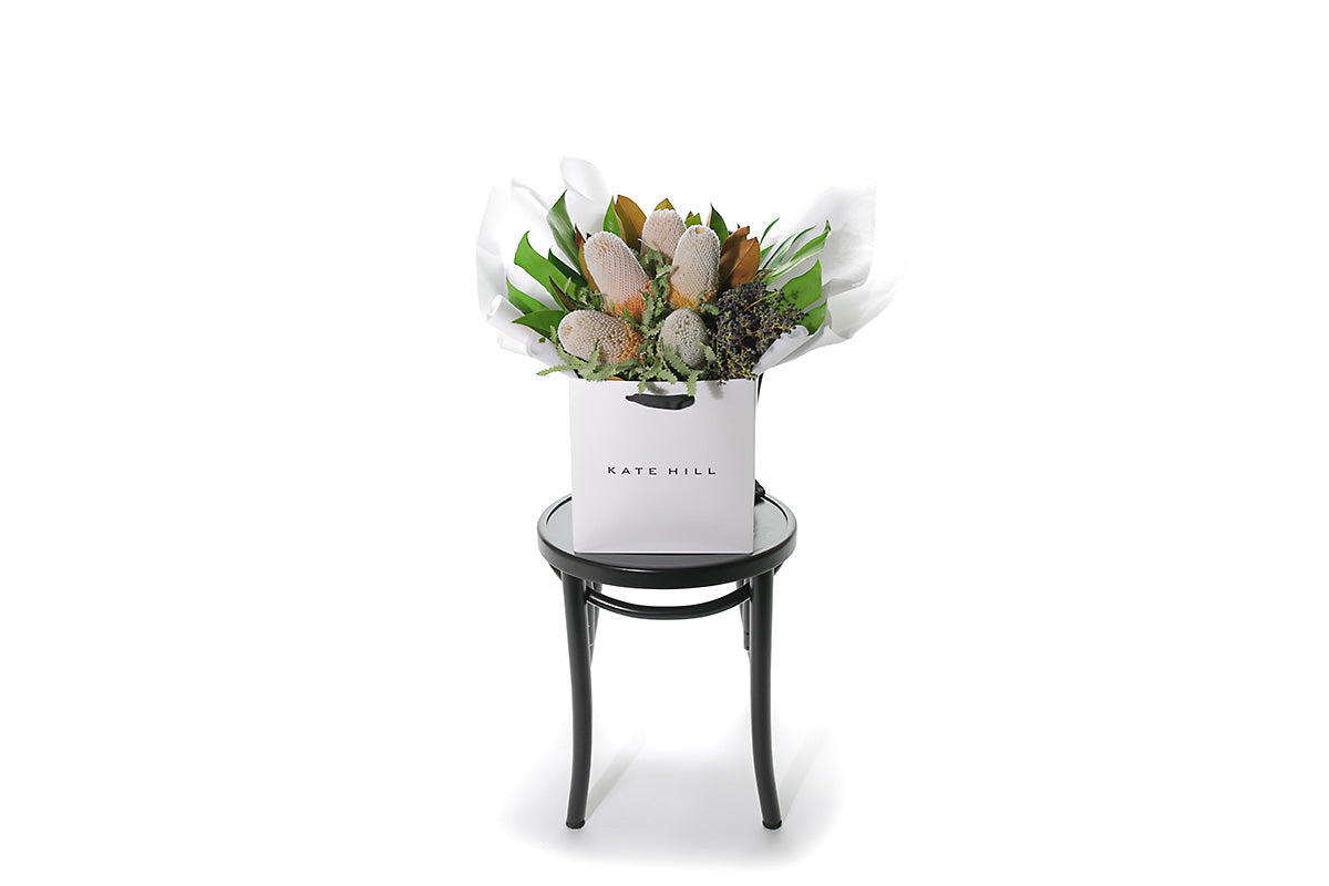 Wide image of A seasonal native bouquet pictured with banksias and seasonal foliage. Bouquet sitting on a black bentwood chair with white background.