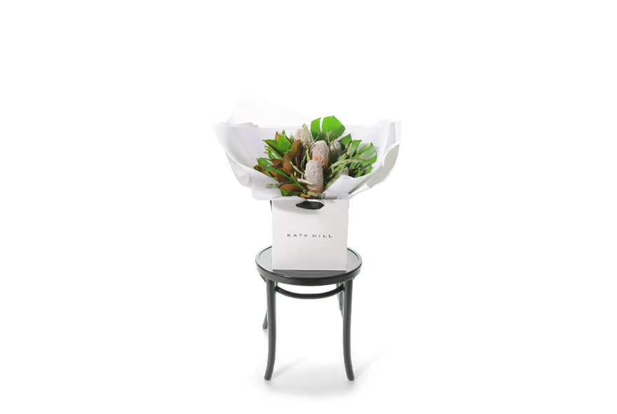 A seasonal Australian Native flower bouquet designed with native foliages, banksias (or seaosnal native flowers) and lush green, brown foliages. Medium to large in size. Presented beautifully in the signature Kate Hill Flowers style in the flower bag. Bouquet bag is sitting on a black bentwood chair and the view is from a far so you can see the honest size of the flower bouquet.