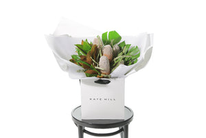 A seasonal Australian Native flower bouquet designed with native foliages, banksias (or seaosnal native flowers) and lush green, brown foliages. Medium to large in size. Presented beautifully in the signature Kate Hill Flowers style in the flower bag. Bouquet bag is sitting on a black bentwood chair.