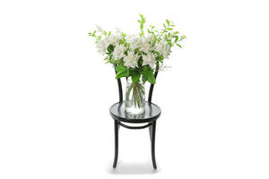 White reflexed roses displayed in a clear glass tapered vase sitting on a black bentwood chair with white background. Wide image of vase design. 