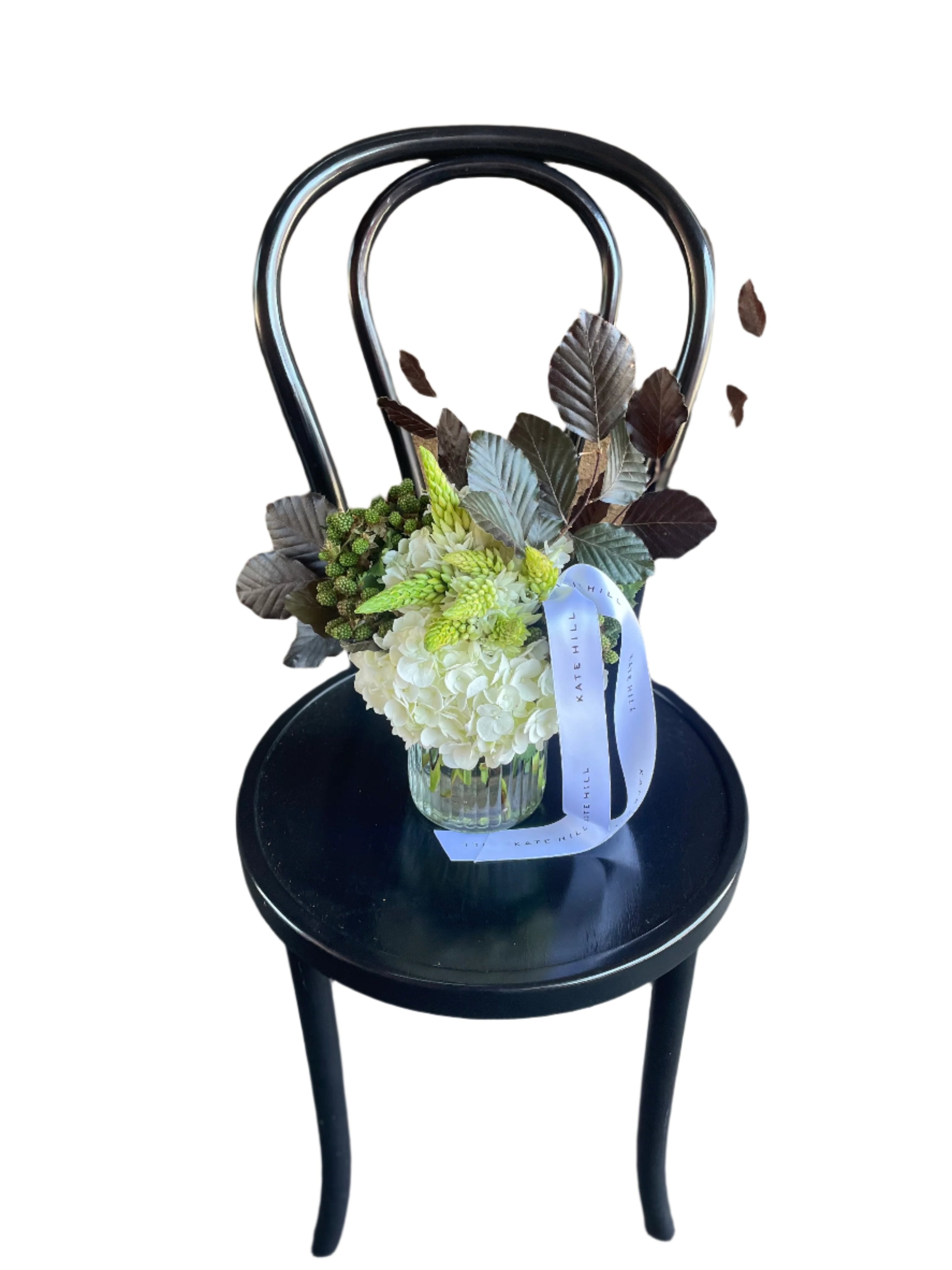 Image from above of a small ribbed vase featuring seasonal white and green christmas flowers. Vase design sitting on black bentwood chair with white wall background.