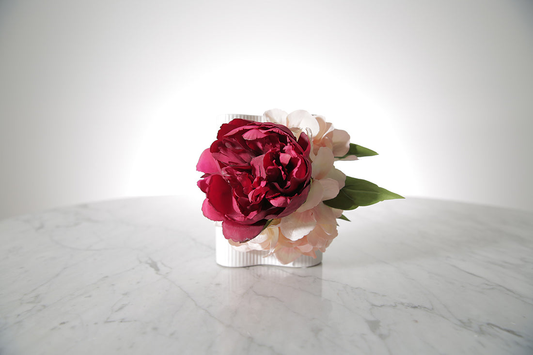 Kate Hill Flowers artificial burgundy peony and blush hydrangea wrist corsage on silver elastic wristlet displayed on a marble table with a white background.