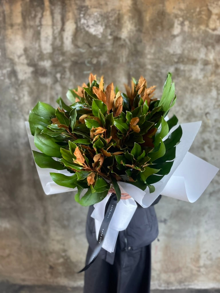 Florist holding a A large bouquet of magnolia foliage and monsteria leaves. Florist standing in front of white wall.