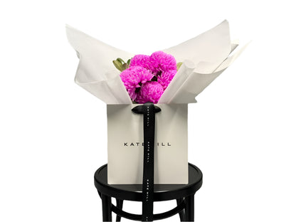 Bouquet of hot pink flowers in a white paper wrap sitting on a black wood chair, against a white wall.