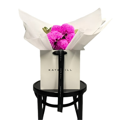 Bouquet of hot pink flowers in a white flower bag sitting on a black bentwood chair with a white wall background.