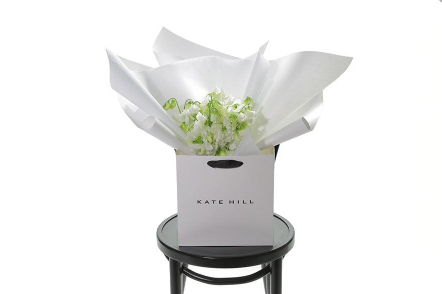 Fragrant bouquet of white sweetpea sitting on a black bentwood chair. Bouquet is beautifully wrapped in the Kate Hill signature wrapping and placed into a Kate Hill flower bag. Our CAMILA Sweetpea Flower Bouquet is an all rounded, simple dome posy featuring fragrant White Sweet-pea. A petite, sweet, fragrant posy perfect for those who love elegant fragrance and who appreciate Sweet-peas for their soft beauty and scent.