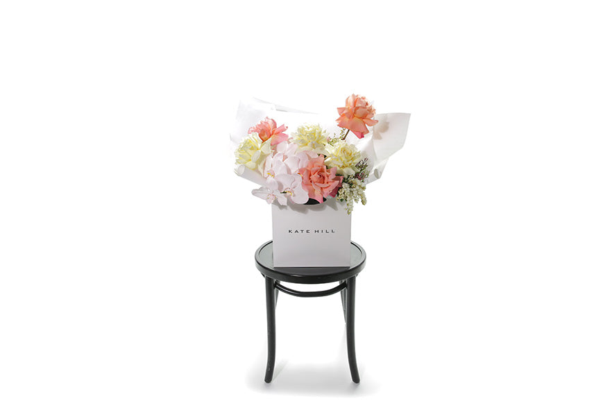 Wide image of the Charlie bouquet. Soft pastel bouquet in lemon, ivory, blush and watermelon tones. Bouquet is wrapped beautifully in white signature wrapping and sitting in kate hill flower bag. Bouquet bag is sitting on a black bentwood chair with white background.