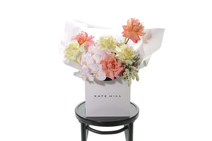 Soft pastel bouquet in lemon, ivory, blush and watermelon tones. Bouquet is wrapped beautifully in white signature wrapping and sitting in kate hill flower bag. Bouquet bag is sitting on a black bentwood chair with white background.