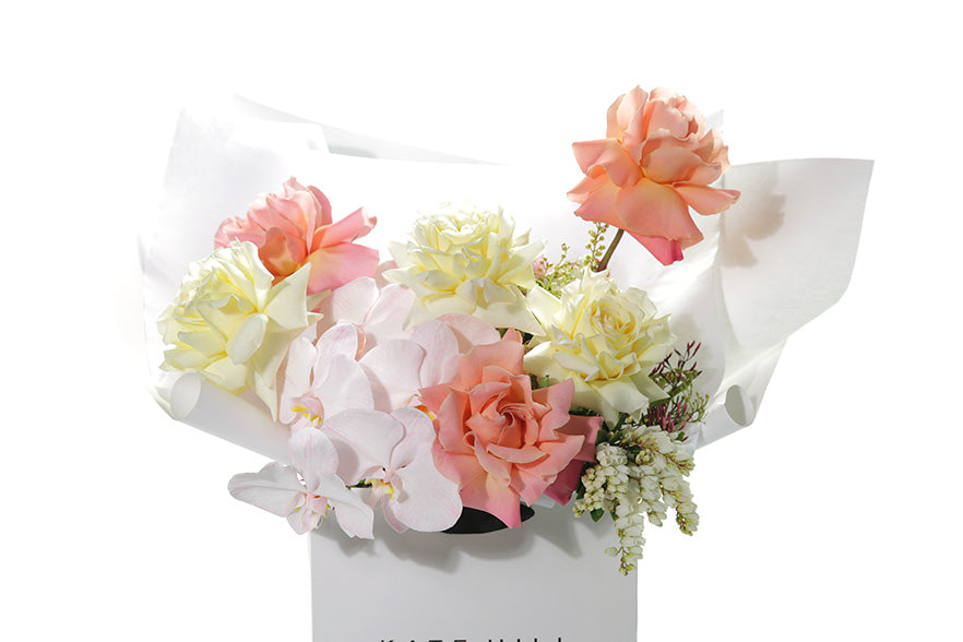 Close up image of the Charlie bouquet. Soft pastel bouquet in lemon, ivory, blush and watermelon tones. Bouquet is wrapped beautifully in white signature wrapping and sitting in kate hill flower bag. Bouquet bag is sitting on a black bentwood chair with white background.