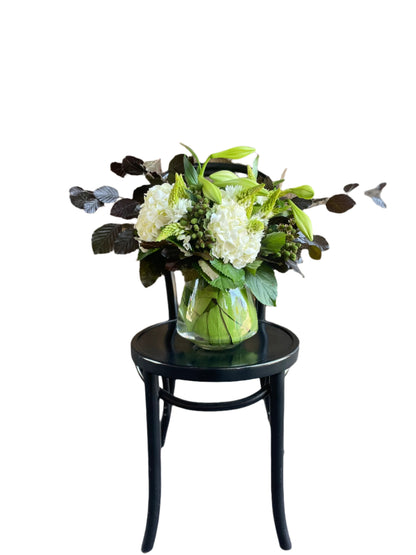 A christmas flower vase design displaying white and green festive flowers sitting on a black bentwood chair with a white background.