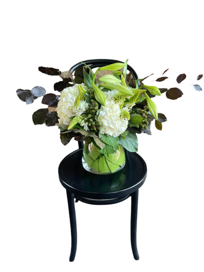 A christmas flower vase design displaying white and green festive flowers sitting on a black bentwood chair with a white background.