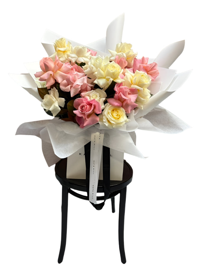 Large bouquet of mixed pastel roses including pink, watermelon, buttery lemon and white roses, sitting on a small black stool against a white wall.