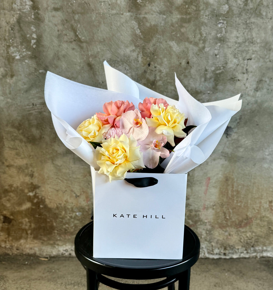 Close up image of A medium sized lemon and blush guft bouquet in kate hill flower bag, sitting on a black bentwood chair in front of a concrete wall.