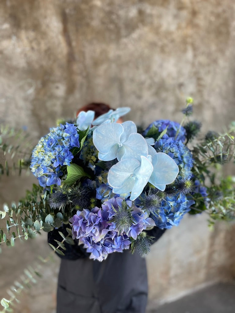 Close up image of Florist holding a table hedge of blue seasonal flowers and grey blue foliages. Florist standing in front of a concrete wall.