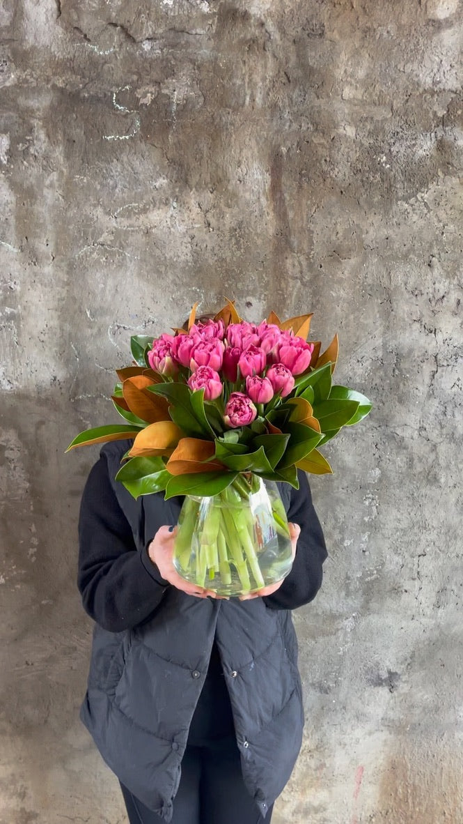 Wide angle of Florist holding a clear tapered vase full of pink double tulips and magnolia foliage, in front of a concrete wall.