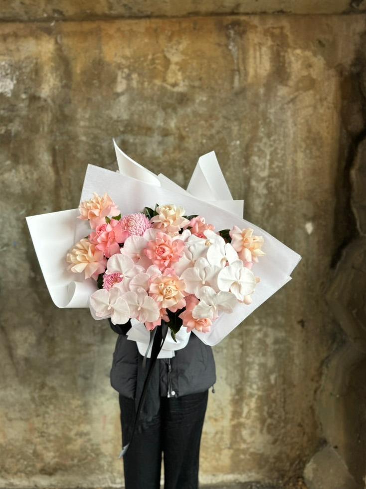 A luxe blush flower bouquet wrapped in signature white wrapping. Florist in black holding the blush flower bouquet in front of a white wall.