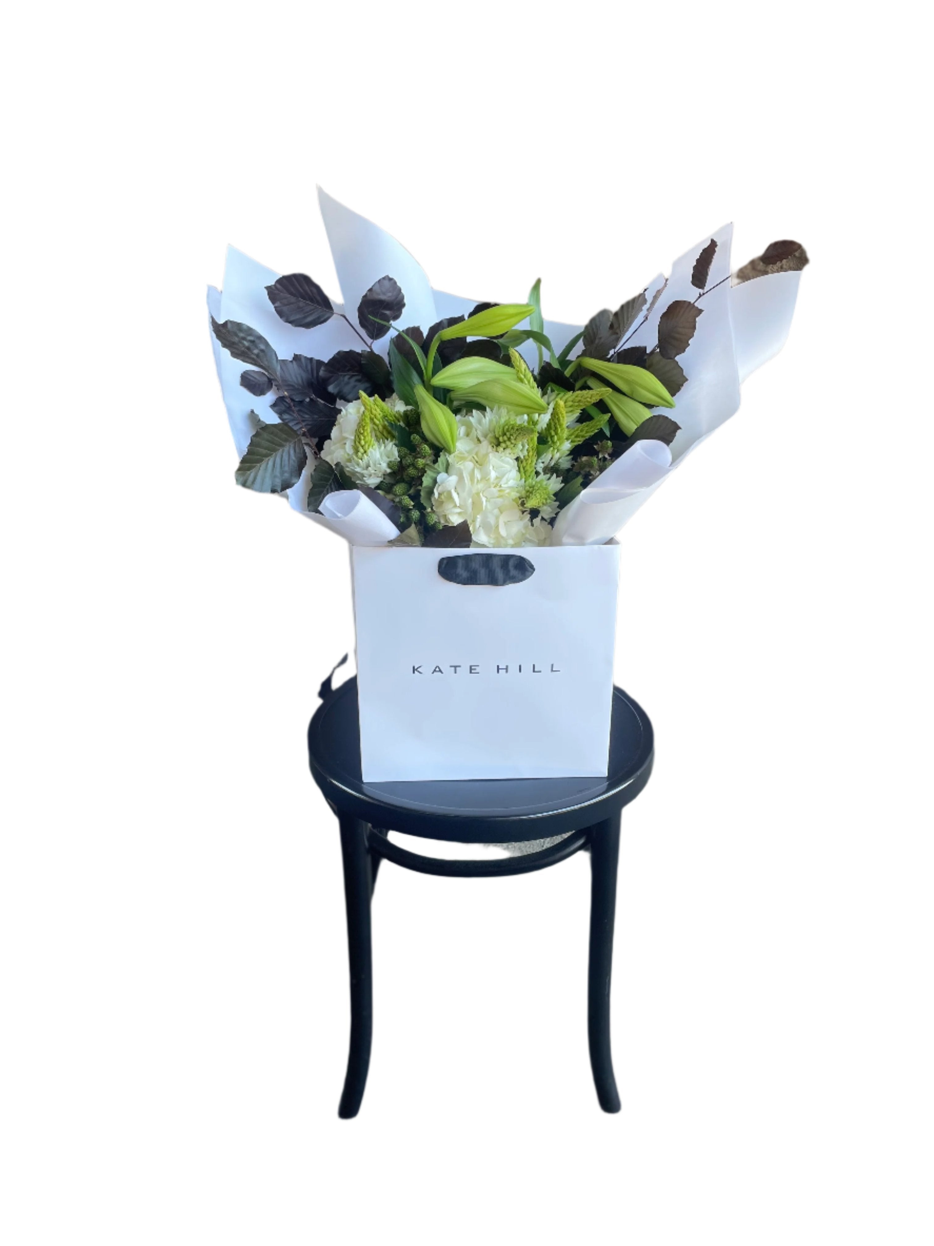 A wide image of our festive white and green bouquet, beautifully wrapped in signature white wrapping sitting on a black bentwood chair with a white wall background.