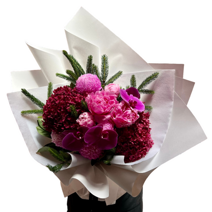 Close up image of a large bouquet of flowers in hot pink, magenta and burgundy colours, detailed with a hint of Christmas spruce foliage. Bouquet is wrapped beautifully in white signature wrapping and florist is standing against a white wall. 