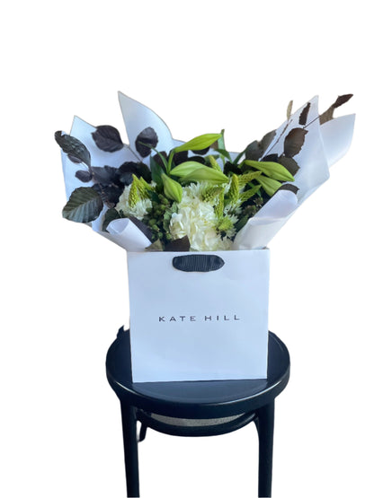 A festive white and green bouquet, beautifully wrapped in signature white wrapping sitting on a black bentwood chair with a white wall background.