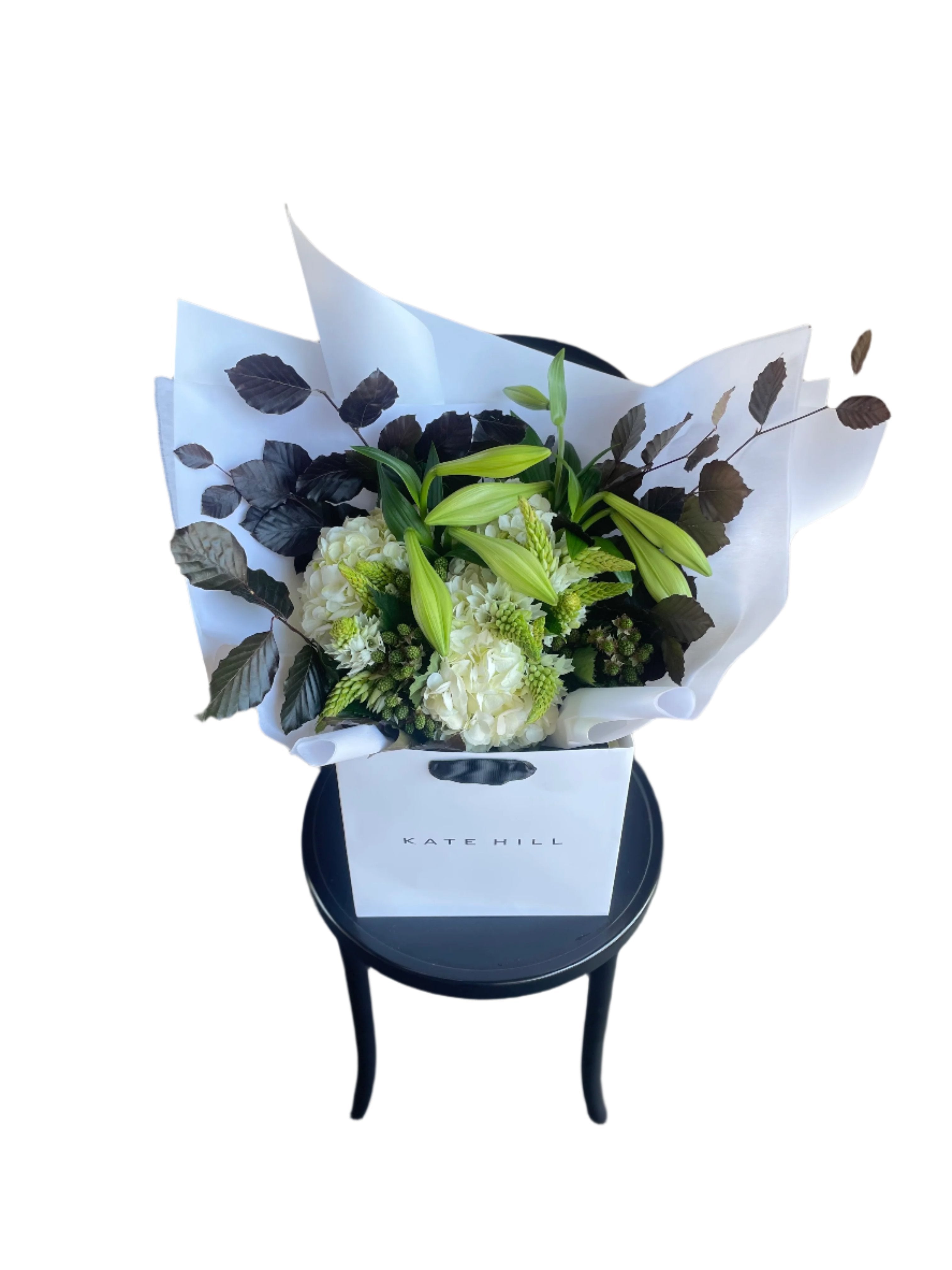 A festive white and green bouquet, beautifully wrapped in signature white wrapping sitting on a black bentwood chair with a white wall background.